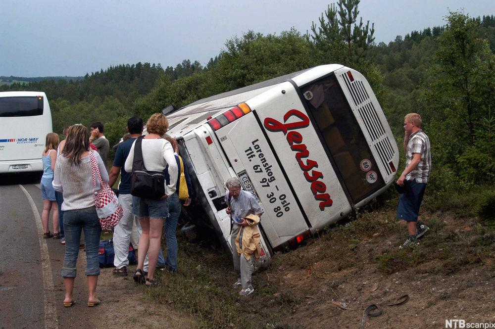 Ein buss ligg velta i ei grøft. Utanfor står evakuerte passasjerar med tilgrisa klede. Foto.