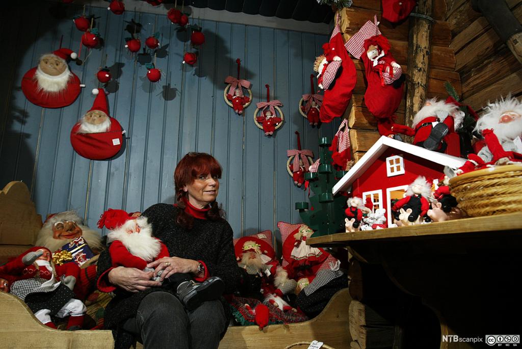 Eva Johansen sitter på en benk i et blåmalt rom. Veggene er fulle av julenisser og annen juledekorasjon. Hun holder i en julenisse. Foto.