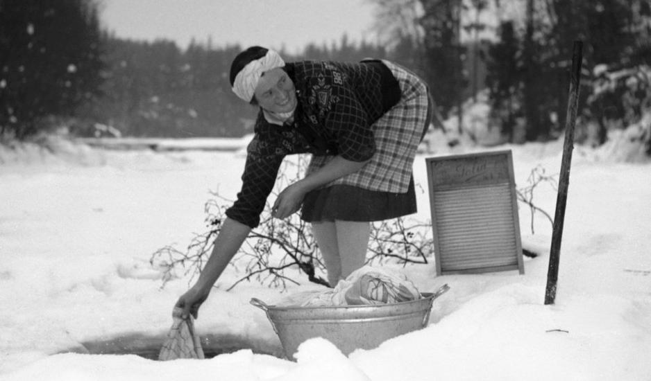 Kvinne vasker klær i et hull i isen i en bekk. Foto.