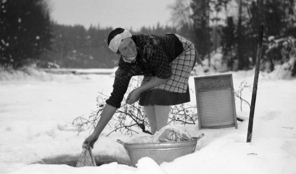 Kvinne vaskar klede i eit hol i isen i ein bekk. Foto.