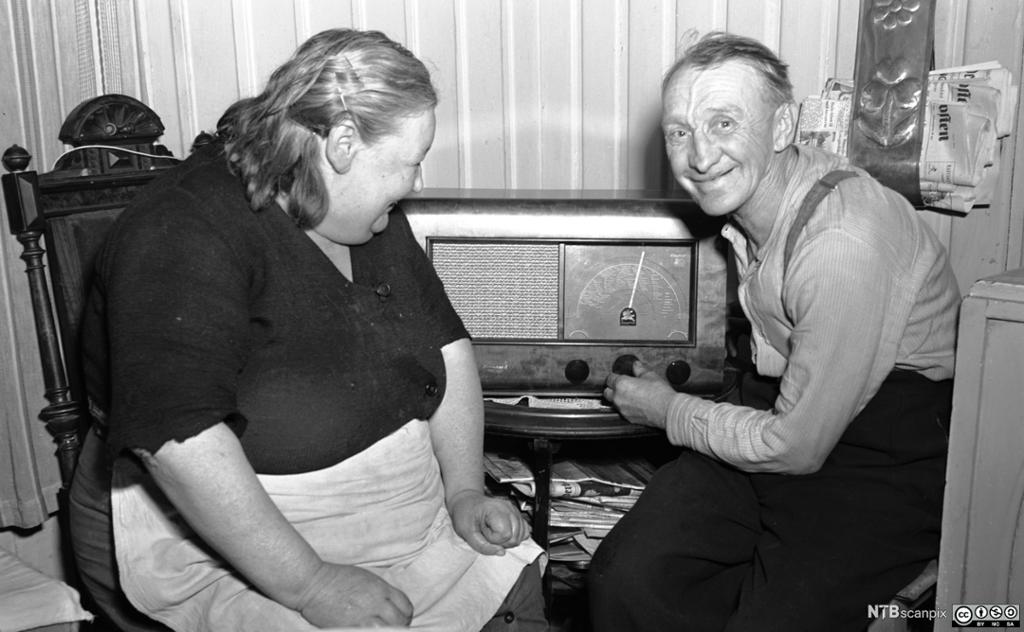 Mann og kvinne sitter foran radioapparat i en typisk 1950-tallstue. De har på seg hverdagsklær, blant annet har hun forkle. Foto.