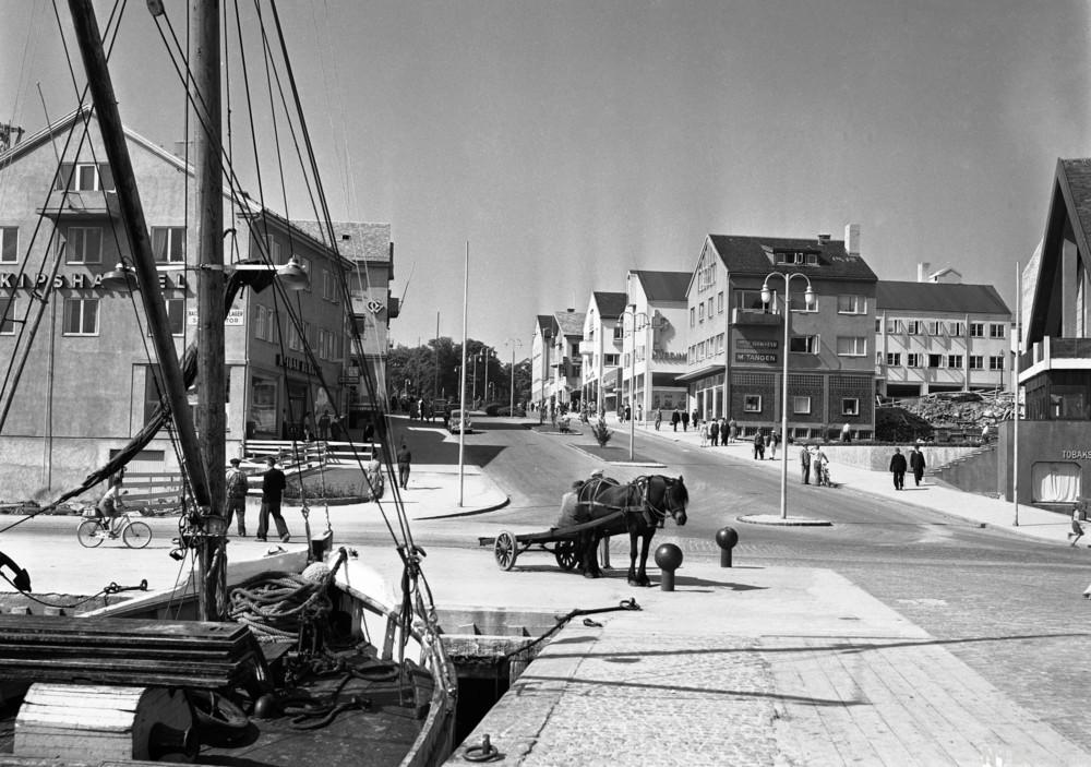 Historisk bilde fra bymiljø med seilbåt ved brygge og hest med kjerre i gaten.foto.