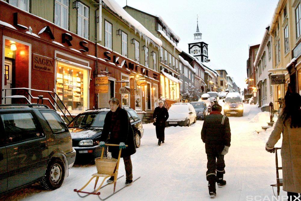 Folk og biler i en snødekket gate med butikker i Røros sentrum. Foto.