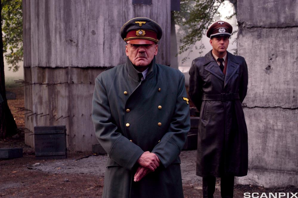 To menn i militæruniform står foran en bunker. Den forreste har smal, mørk bart og ser svært lite fornøyd ut. Foto.