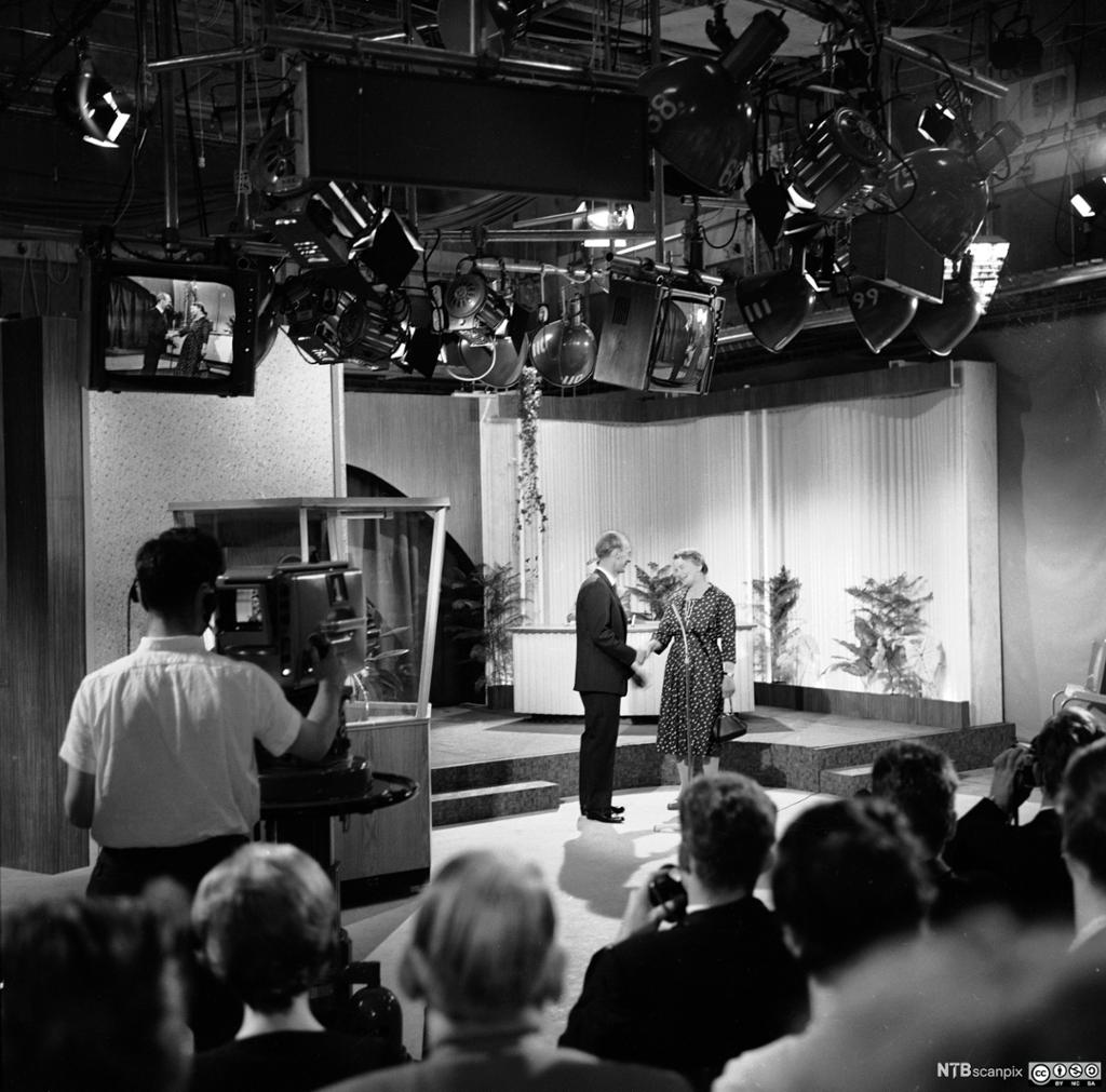 Foto av innspeling av fjernsynsprogram i eit studio med publikum. Ein mann og ei kvinne handhelsar på scenen.
