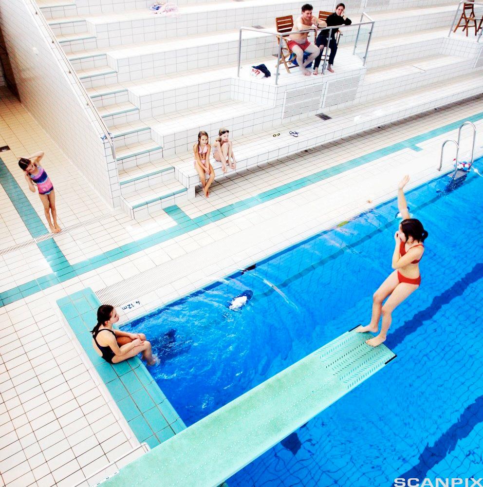 Ei jente hopper baklengs fra et stupebrett i en svømmehall mens andre unge mennesker ser på. Foto.