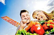 En mann og en kvinne knasker gulrøtter bak en haug grønnsaker, med blå himmel og logoen til VGs Vektklubb i bakgrunnen. Foto.