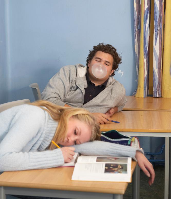 To ungdommer, en gutt og ei jente, sitter ved skolepulter. Gutten blåser ei tyggisboble, og jenta ligger slapt utover pulten. Foto.