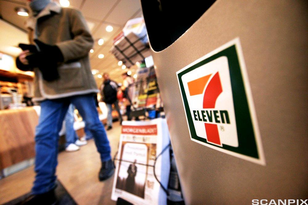 Foto tatt inne i en 7-Eleven-kiosk, med disken i bakgrunnen, kunder rundt omkring i butikken og en stor flate med 7-Eleven-logoen fremst i bildet.