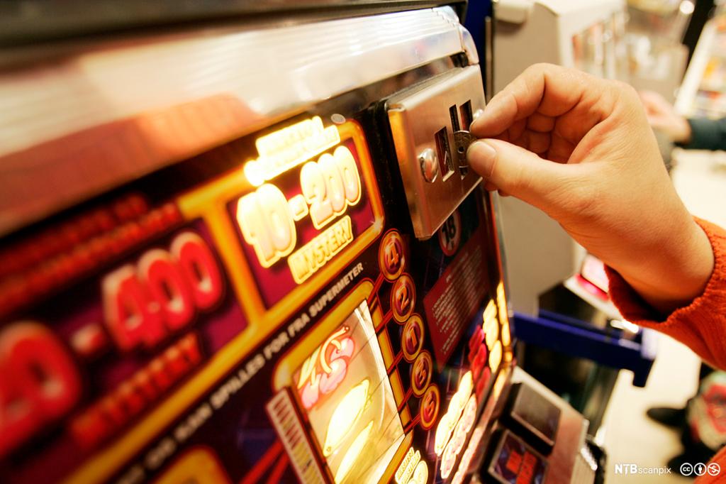 Nærbilde av spilleautomat. Ei hand som putter på en mynt. Foto.