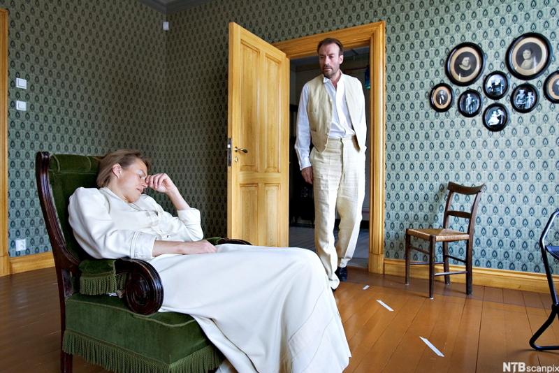 Scene fra Et dukkehjem av Henrik Ibsen. Nora sitter på en stol og ser fortvila ut. Thorvald er på vei inn i stua. Foto. 