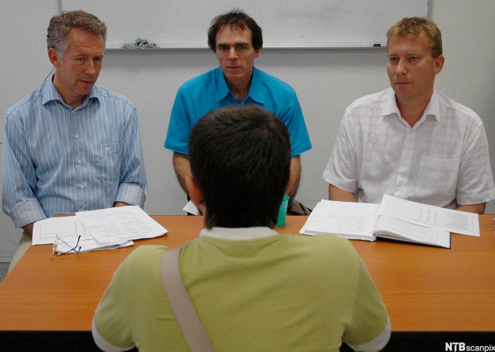 Ein mann med ryggen til snakkar med tre menn på den andre sida av eit bord. Dei tre mennene har papir framfor seg. Foto.