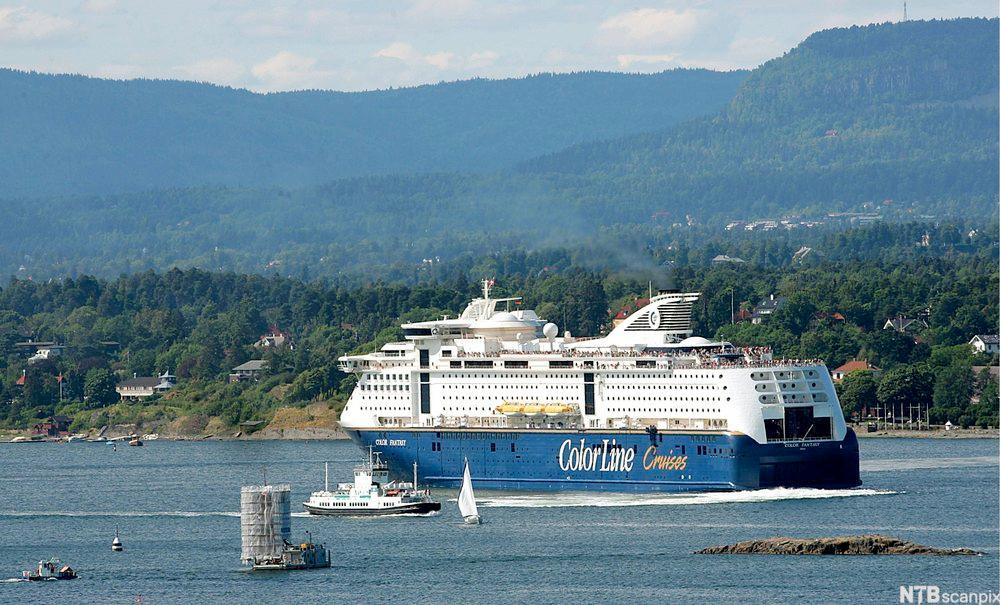 Ei stor hvit og blå ferge merket Color Line Cruises passerer andre fartøy i en fjord. I bakgrunnen av bildet er et skogkledt landskap med åser og spredt bebyggelse. Foto.
