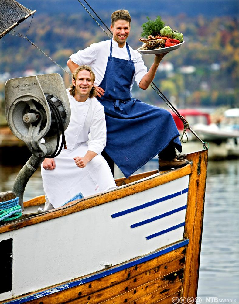 To kokker på en båt. Den ene holder et fat med råvarer. Foto.