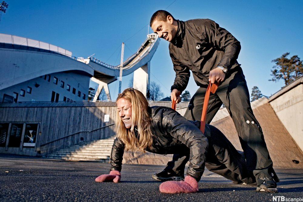 Scene fra fjernsynsprogrammet «Puls». En mann hjelper en kvinne som tar armhevinger, ved å holde henne i et treningsbånd hun har rundt livet. Foto.