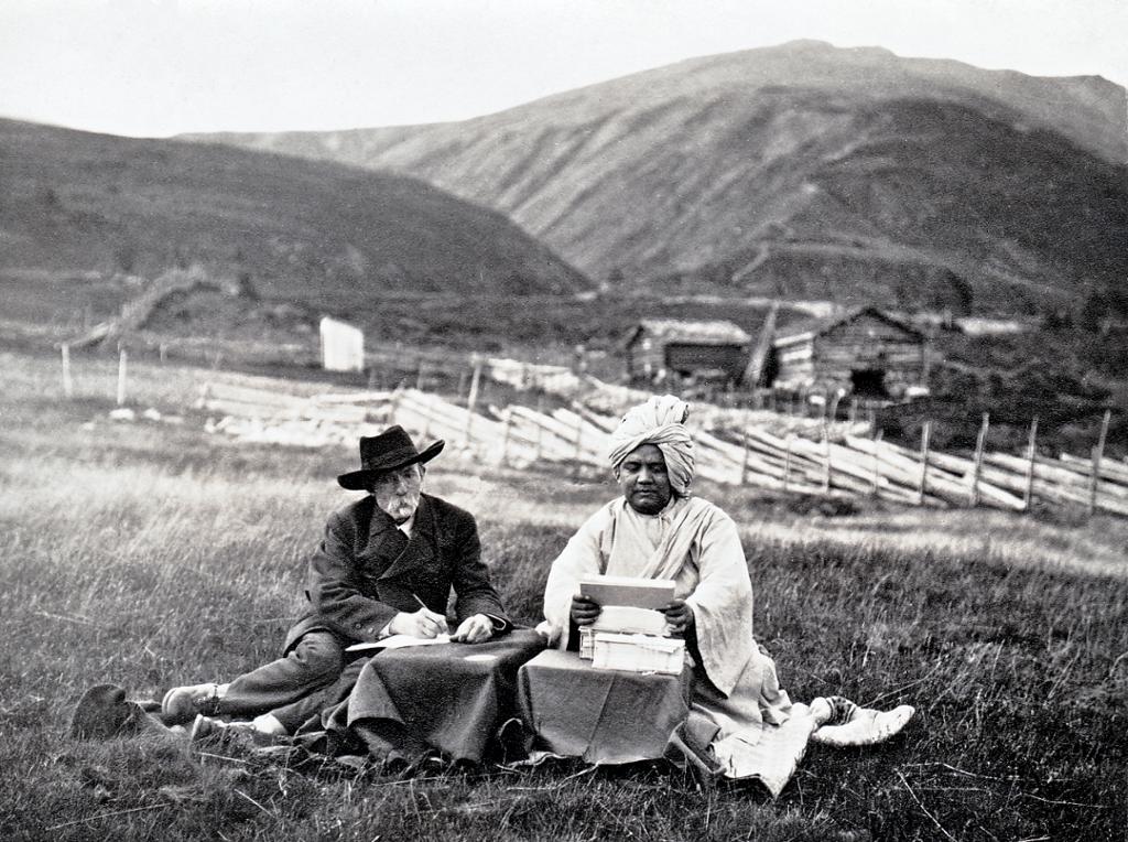 En mann med svart dress og hatt og en mann med turban sitter sammen i gresset ved hvert sitt lille bord. Mannen med hatten skriver og mannen med turbanen leser. I bakgrunnen er det fjell, seterbygninger og en skigard. Svart-hvitt. Foto.