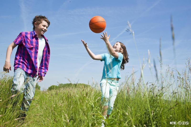 To barn, en gutt og ei jente, løper i ei eng og kaster en rød ball mellom seg. Foto.