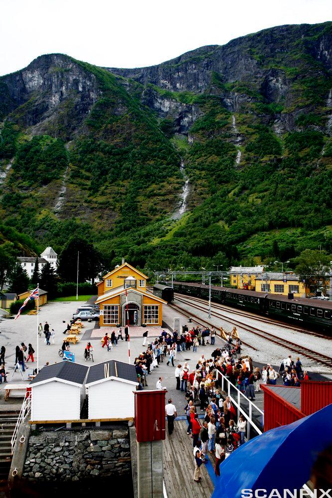 Jernbanestasjon med høye fjell i bakgrunnen. Mange turister står i kø og venter på toget. Foto.