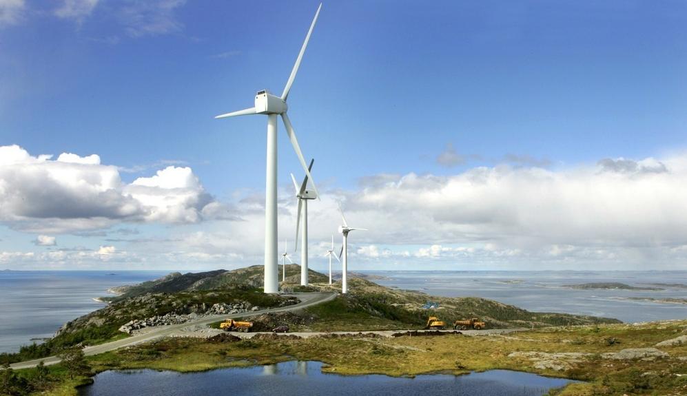 Fem vindmøller fra Hundhammerfjellet vindmøllepark står i terrenget ute ved kysten. Det er et ferskvann i forkant av vindmøllene. Foto.