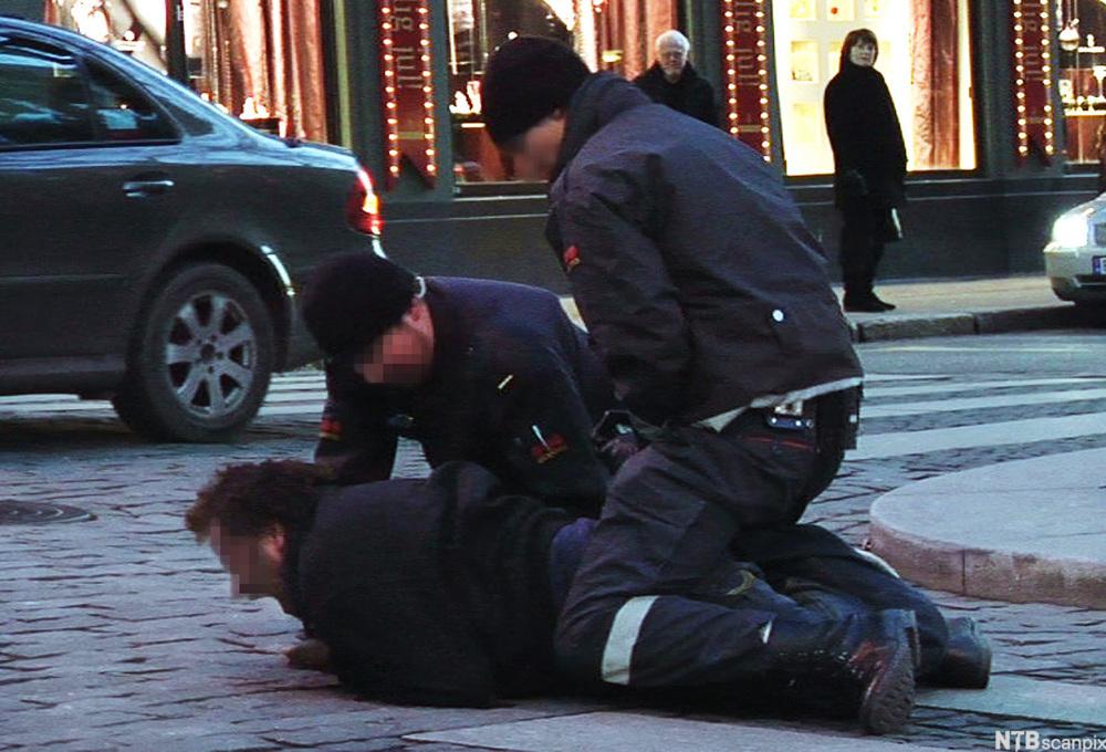 Arrestasjon der ein person ligg på magen på bakken og blir halden nede av to personar i vektaruniform. Foto.