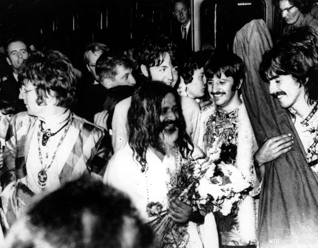 Indisk mann med stort hår og skjegg omgitt av medlemmane av The Beatles. Foto.