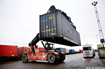 En stor truck løfter en konteiner på en terminal. Like ved står en trailer klar til pålessing. Foto.