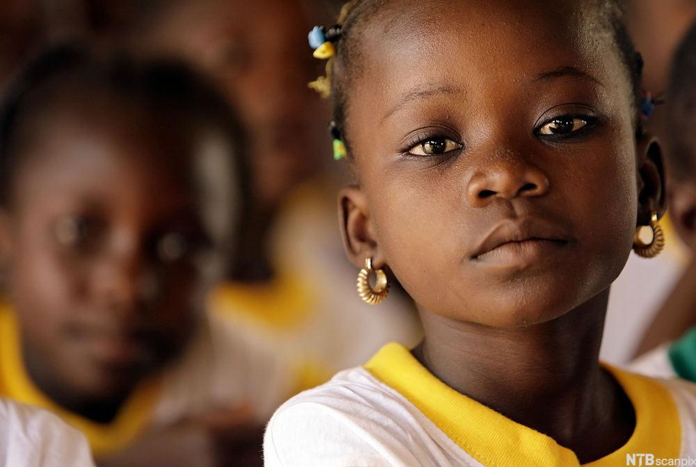 Jente på skole i Burkina Faso. Hun ser i kamera og har skoleuniform på. I bakgrunnen er det flere elever. Foto.