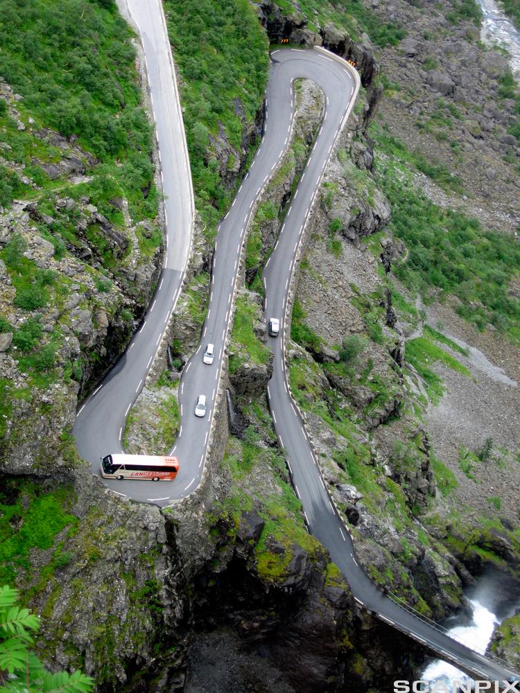 Eit bilete av Trollstigen ovanfrå, som viser krappe svingar oppover fjellsida. Ein buss rundar ein av svingane, og fleire personbilar kjem etter. Foto.