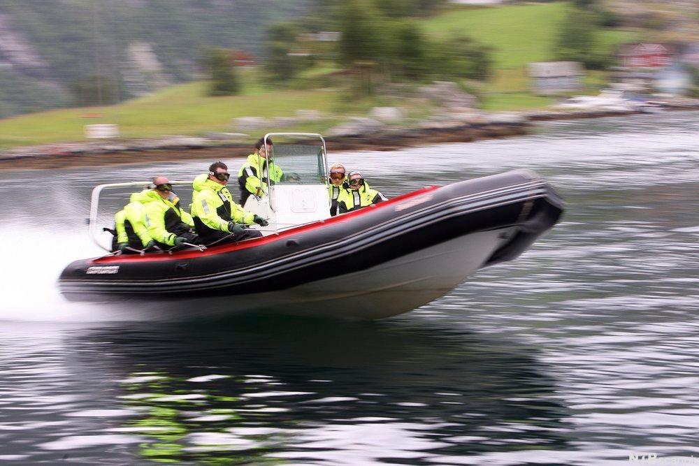 En RIB på vei utover en fjord i høy hastighet. Personene om bord har vernebriller og solide, signalgule jakker. De holder seg fast i et tau langs rekka Foto.