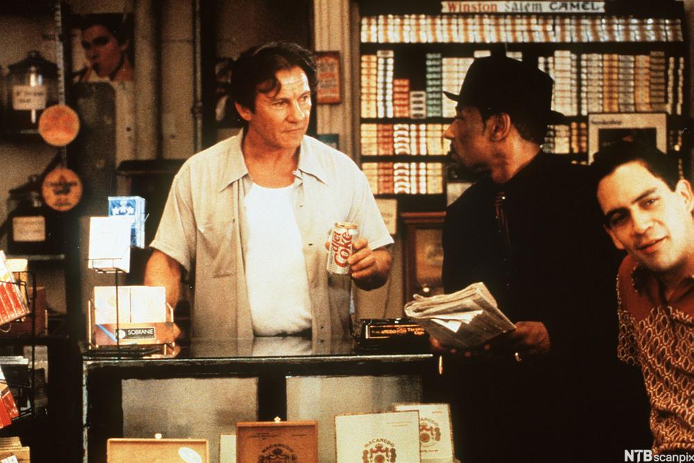 A scene from the film Smoke. Auggie Wren is standing behind the counter of his shop. Two other customers are in the picture. 