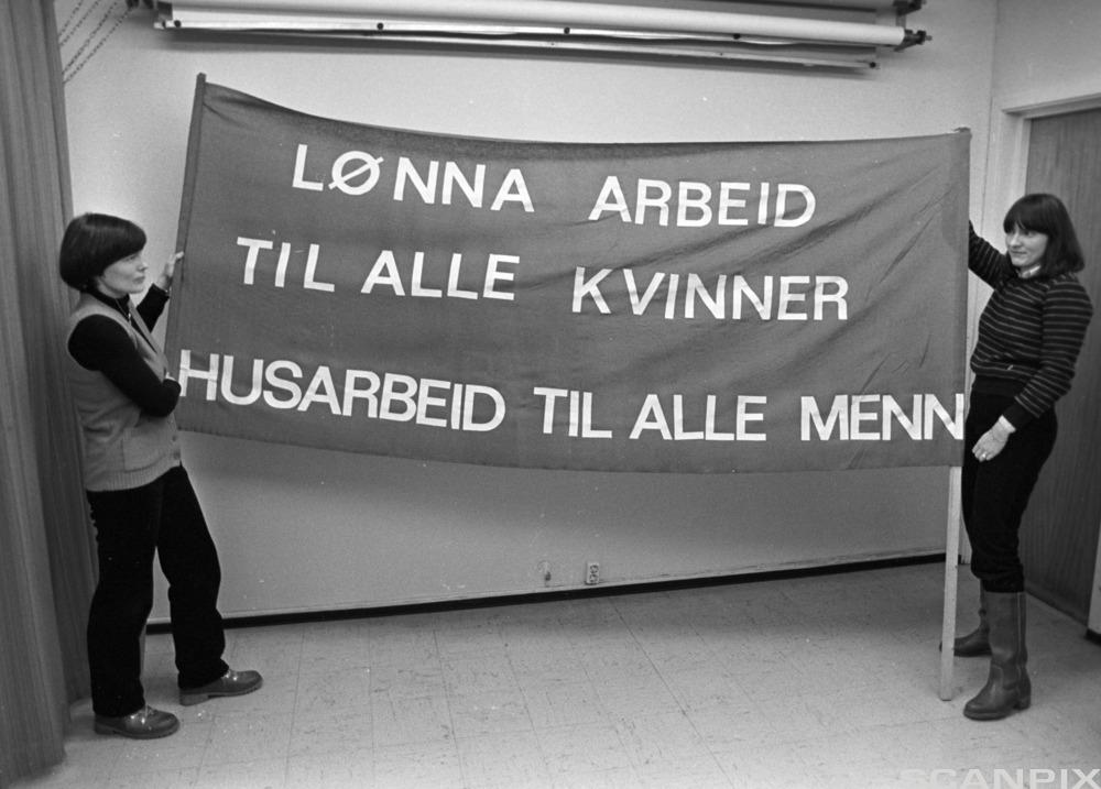 To kvinner som holder en fane med teksten "lønna arbeid til alle kvinner, husarbeid til alle menn". Svart-hvitt-foto.