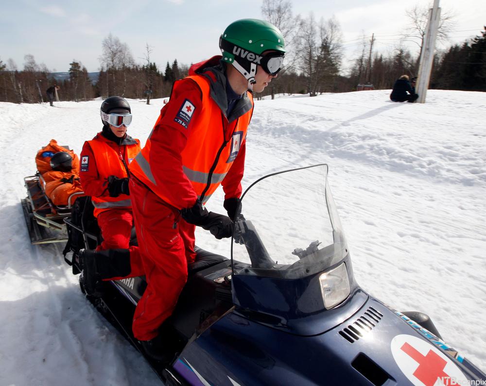 To personar i Røde Kors-uniform køyrer snøskuter med ein slede. Foto.