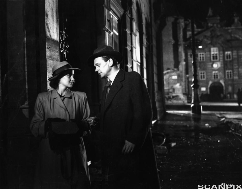Svart og hvitt bilde av kvinne og mann med hatt og frakk i en mørk gate. Foto.