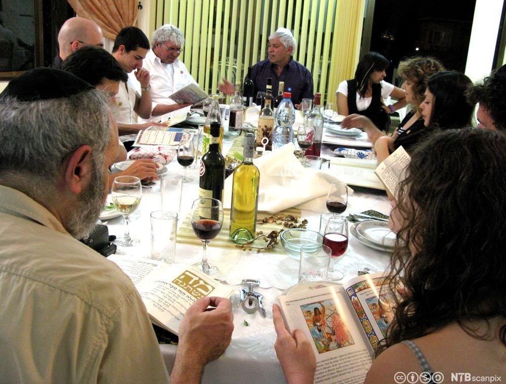 En jødisk storfamilie rundt et festbord med vann og vin. Mange leser i bøker på hebraisk. Foto.