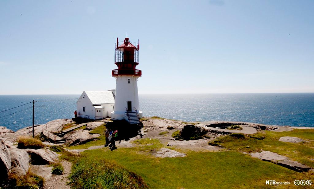 Et fyrtårn og et lite hvitt hus bygd på en klippe. Sjø og blå himmel i bakgrunnen. Foto.