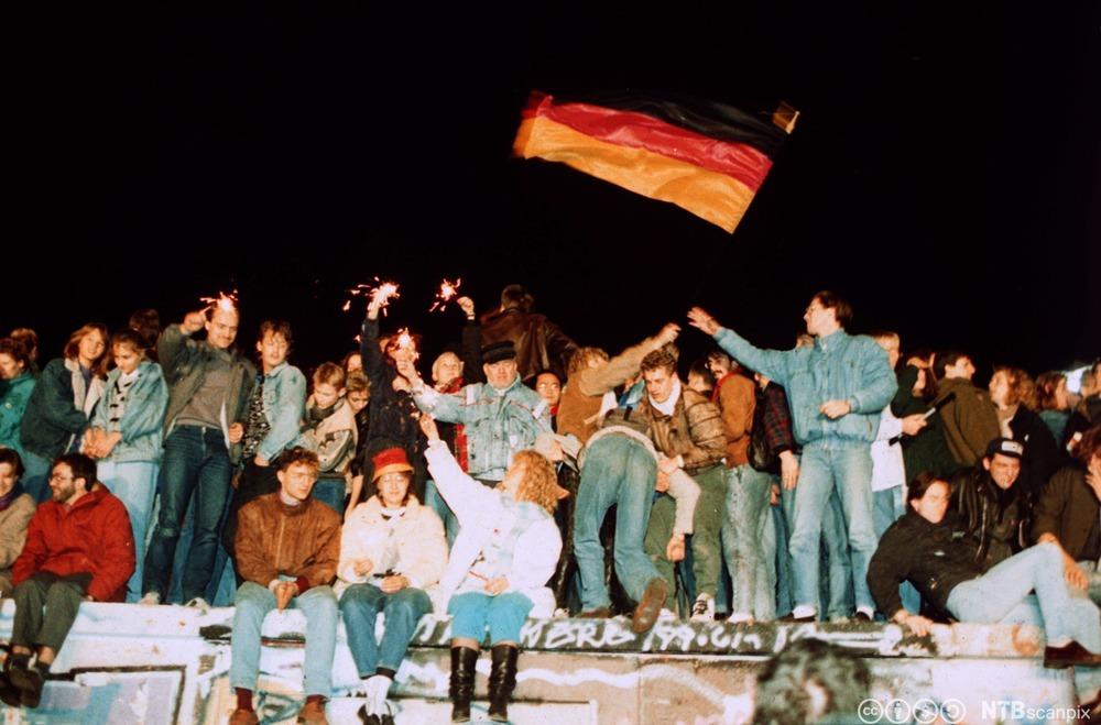 Tett i tett med folk sit og står på Berlinmuren. Dei har stjerneskot og eit stort, vaiande tysk flagg. Foto.