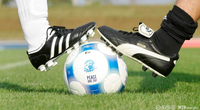 En fotball i midten, en fot i Adidas-sko og en fot i Puma-sko på fotballen. Foto.
