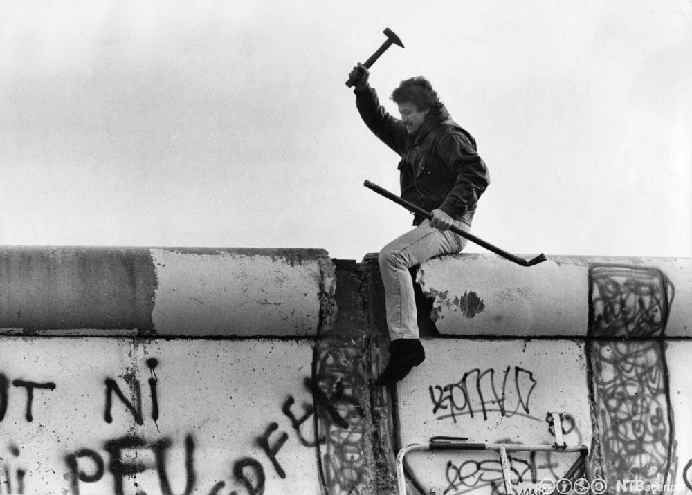 En mann sitter oppe på Berlinmuren og  hakker løs på  den. Foto.