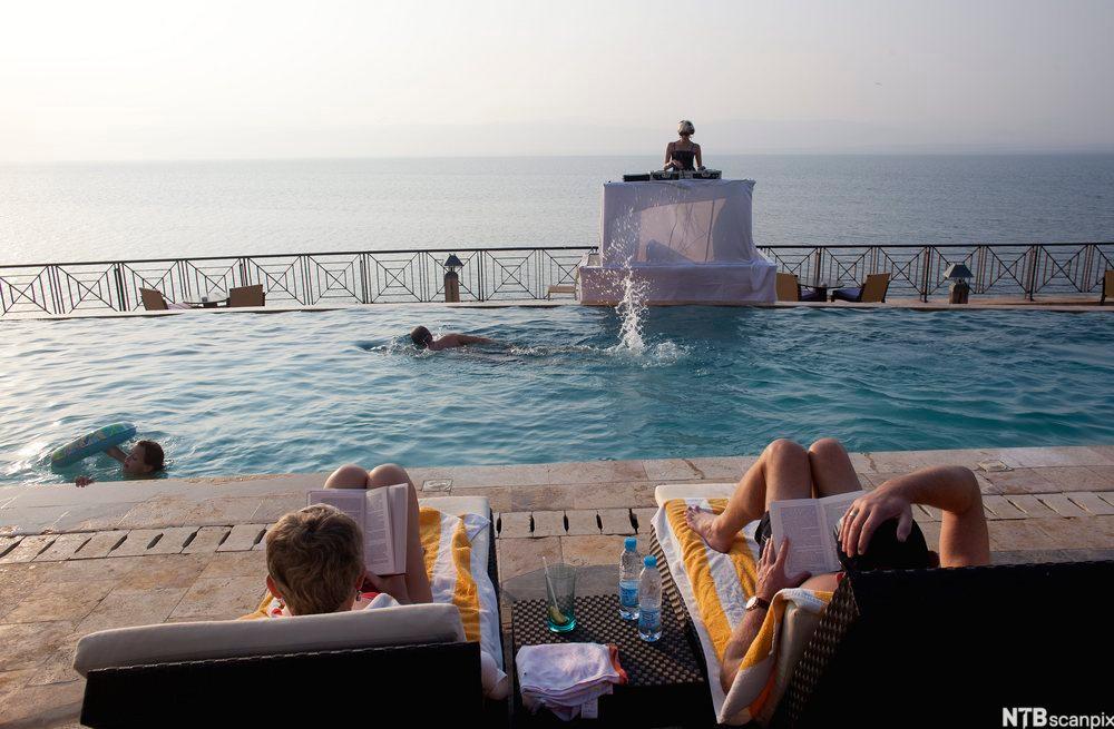 To personer ligger og leser på hver sin solseng ved et badebasseng med utsikt utover havet. Et barn og en voksen bader, og en DJ spiller musikk ved bassenget. Foto. 