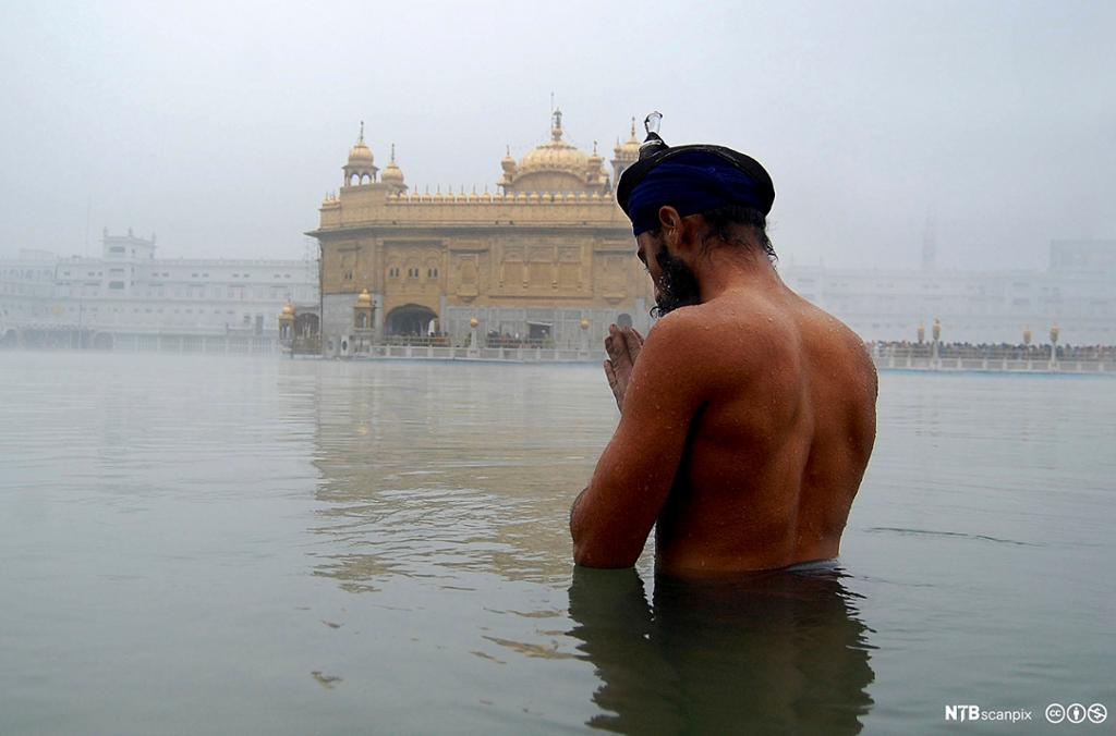 Ein mann står til livet i vatn med turban på hovudet og ber. I bakgrunnen er eit tempel. Foto.