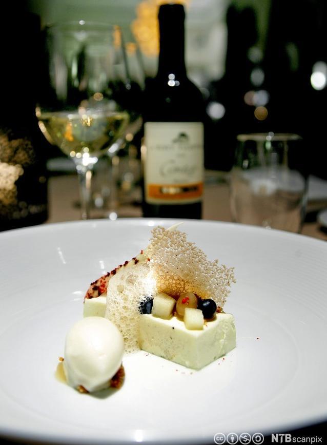 Bilete av ein tallerken med dessert og ei flaske med dessertvin i bakgrunnen. Foto.