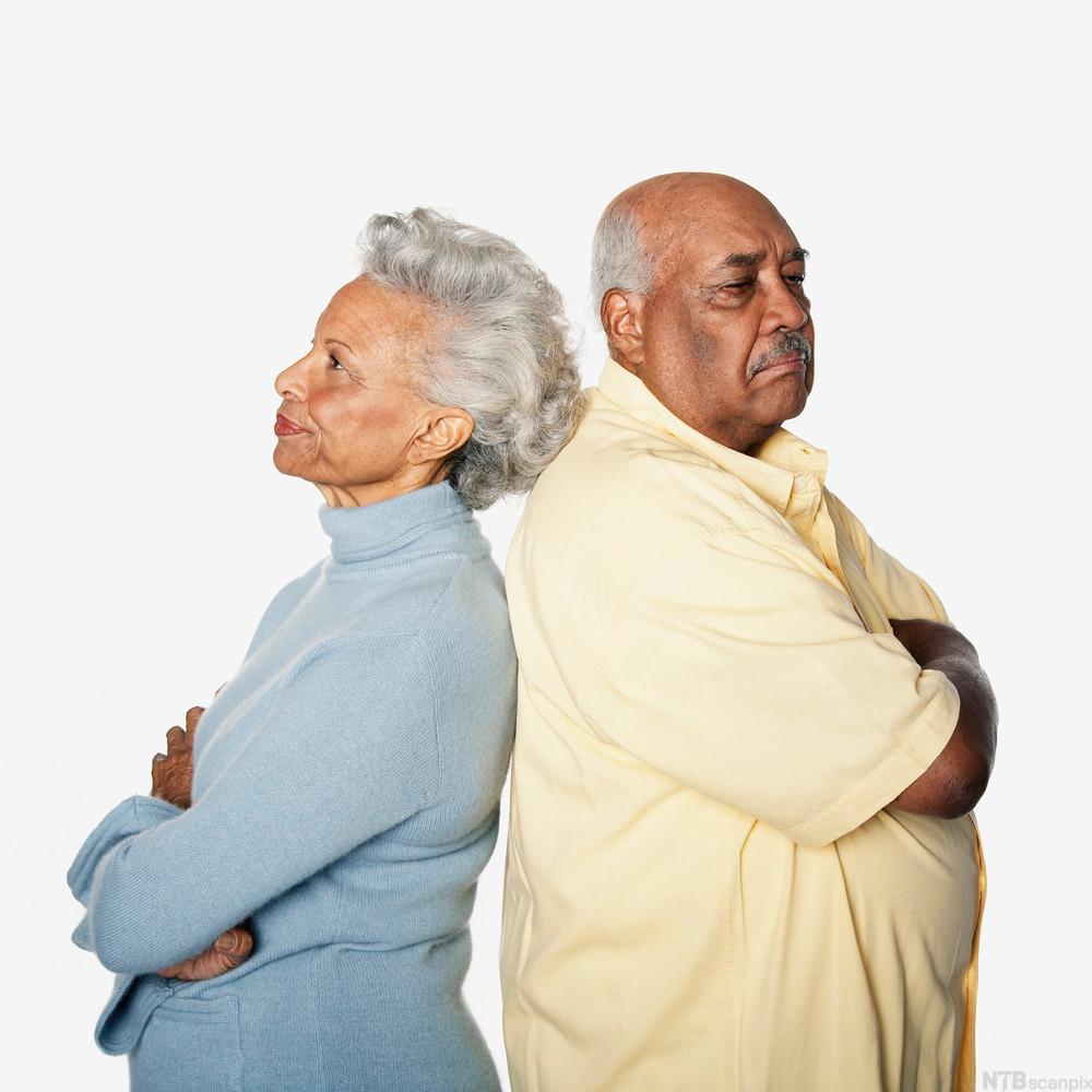 En eldre dame og en eldre mann står med ryggene mot hverandre og armene i kryss. Foto.