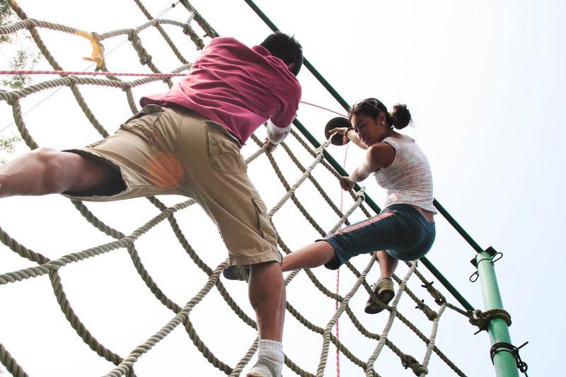 En gutt og ei jente på rundt 16–17 år. De har på seg sommerlige fritidsklær og klatrer i et klatretaustativ. De er to–tre meter over bakken, og jenta har nådd toppen på stativet. Foto. 