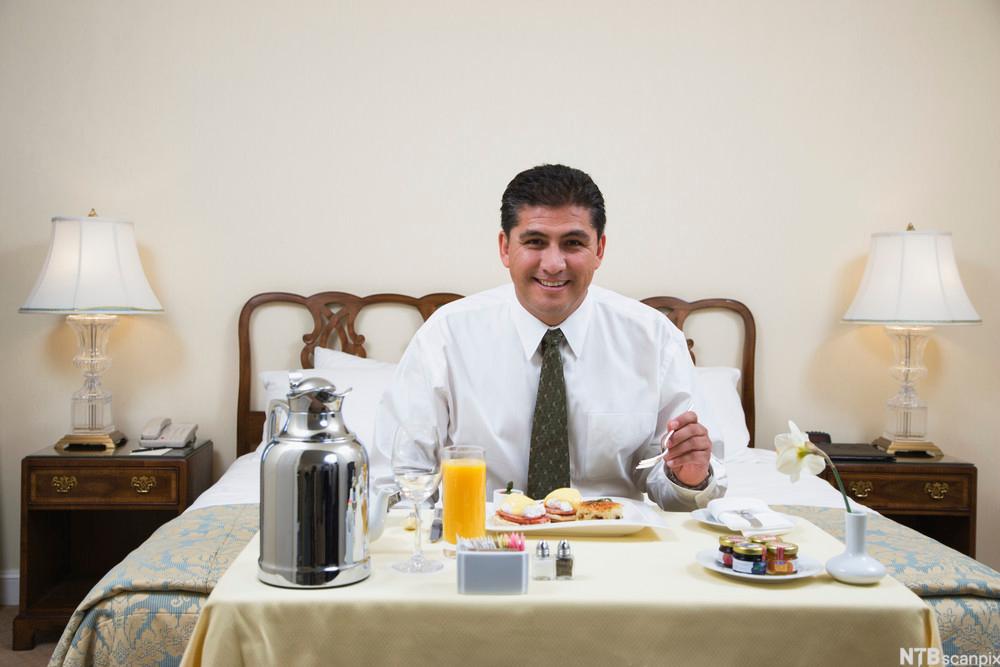 Ein nøgd mann et frukost på hotellrommet, med blant anna kaffi, fersk jus og rundstykke. Foto.
