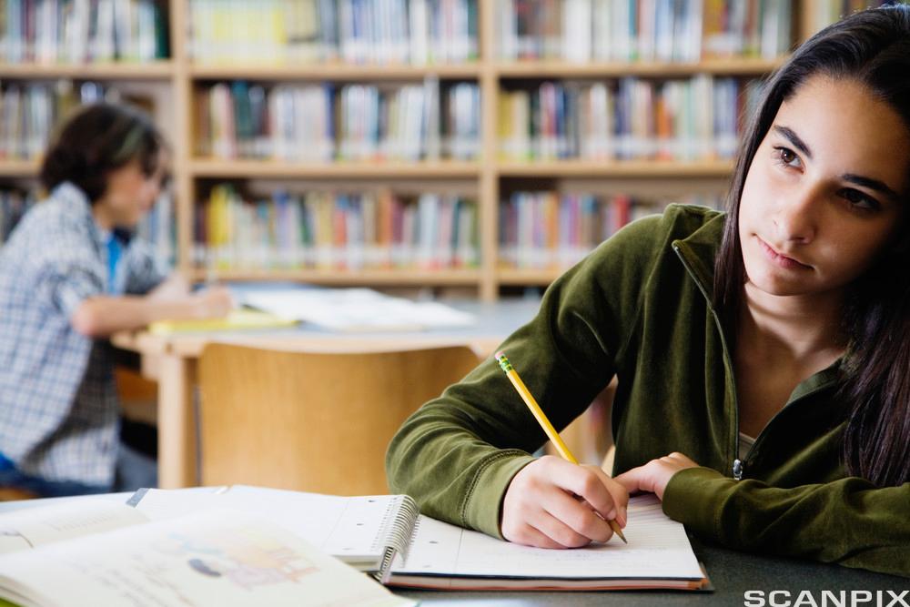 Ei jente sitter på biblioteket med ei skrivebok foran seg på og en blyant i hånda. Hun ser utover rommet. I bakgrunnen ser vi en annen ung person. Foto.