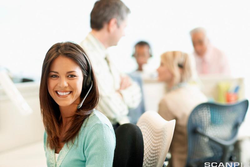 Smilende kvinne med headset sitter på et kontor med kollegaer i bakgrunnen. Foto.