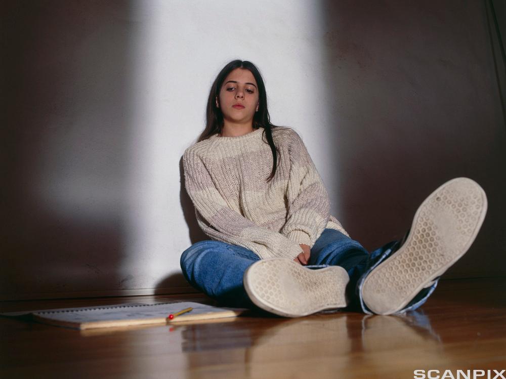 Ung jente sitter på gulvet inntil en vegg og ser nedtrykt ut. Det ligger en notisbok og en penn ved siden av henne på gulvet. Foto.