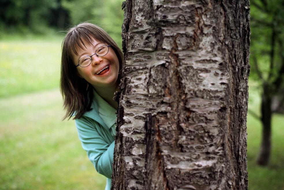Jente med Downs syndrom står bak et tre. Foto.