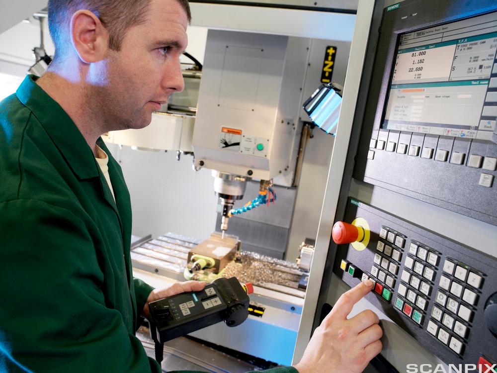 En person som står foran en CNC-maskin og trykker på betjeningspanelet med høyre hånd. Har fjernkontroll til maskinen i venstre hånd. I bakgrunnen et oppspent arbeidsemne på en maskin. Foto.