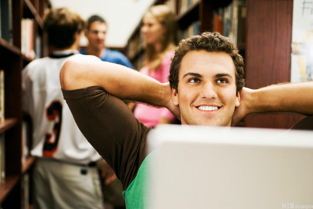 Ung mann sitter bak en dataskjerm med hendene bak hodet og smiler. Foto.